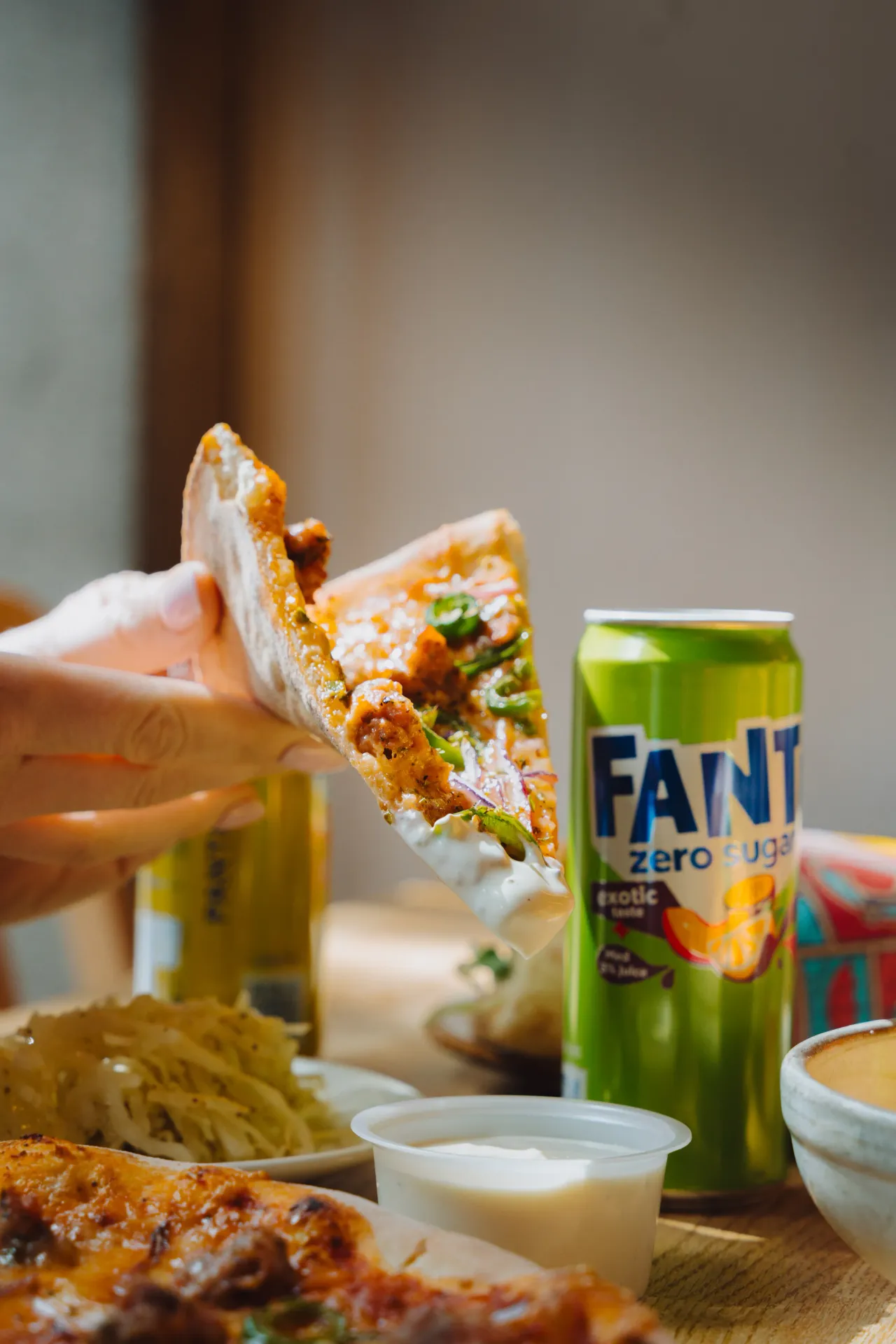 En hand håller en bit pizza över ett bord med en grön burk Fanta Zero Sugar Exotic, fler pizzabitar, sås och en tallrik i bakgrunden.