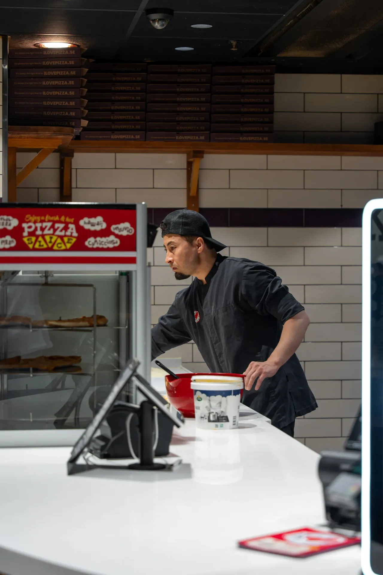 En pizzabagare i svart keps och arbetskläder arbetar bakom disken i en pizzeria med vita kakelväggar och staplade pizzakartonger på hyllor bakom honom.